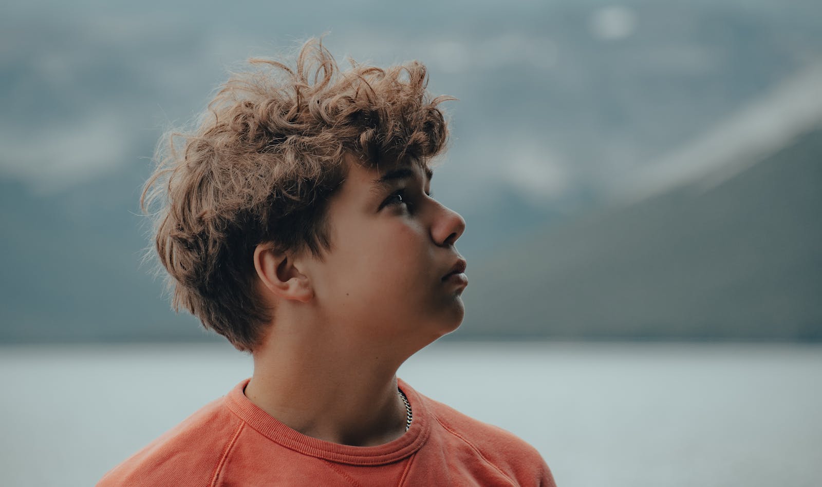 Teenage boy with curly hair in profile view outdoors, thoughtful expression.