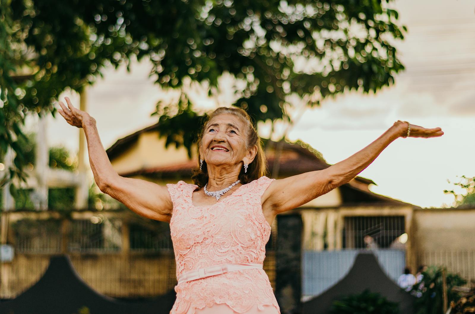 Elderly woman in a pink lace dress smiling and posing outdoors.