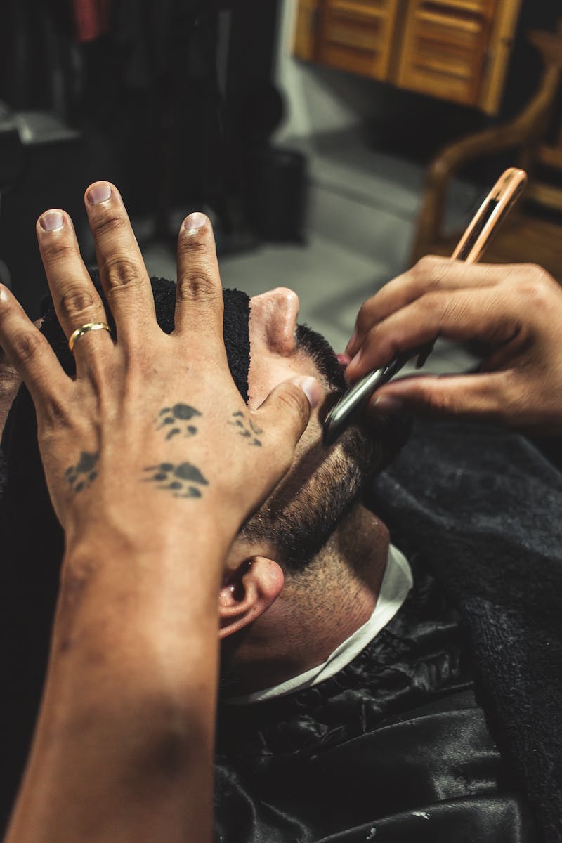 A barber using a straight razor to shave a man's face indoors, showcasing tattooed hands.