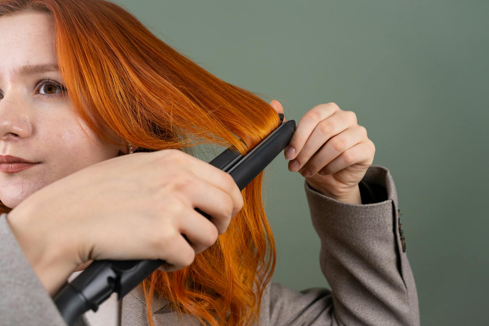 Close-up of a woman styling her red hair using a hair straightener against a green background.