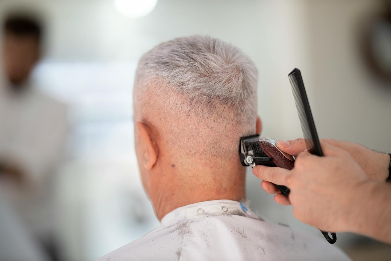 Elderly man getting hair trimmed by barber indoors using clipper and comb.