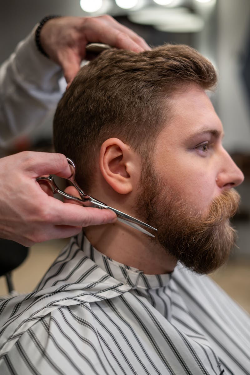 A side view of a barber trimming a bearded man's hair in a barber shop.