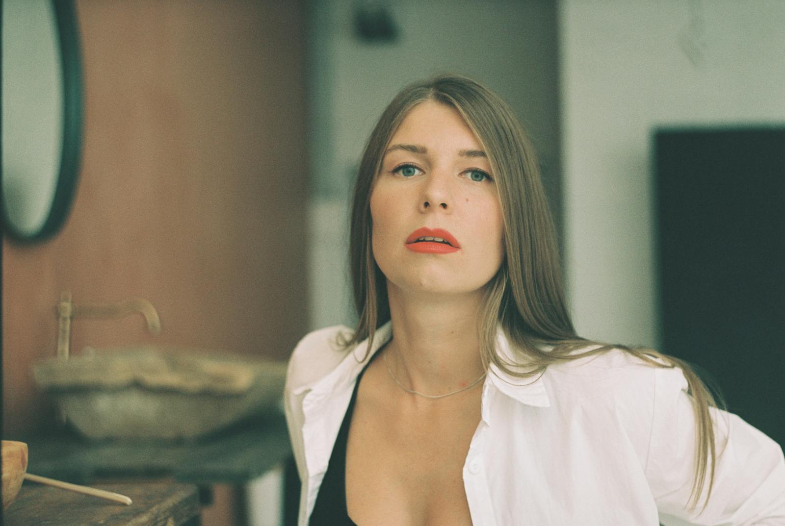 A woman indoors, styled with red lipstick, long hair, and a button-up white shirt.