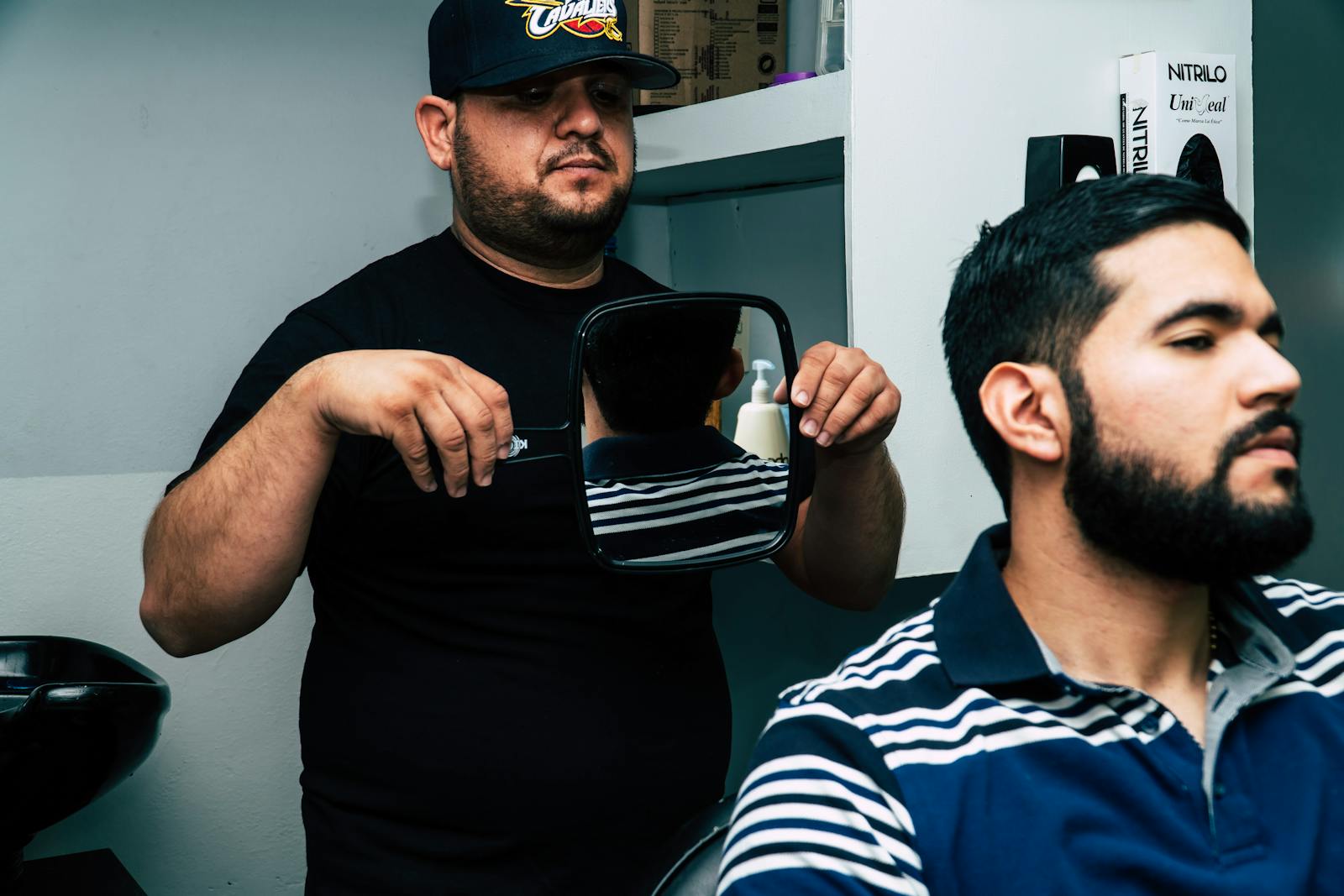 A barber in a cap shows a mirror to a client with a styled beard in a modern barbershop.