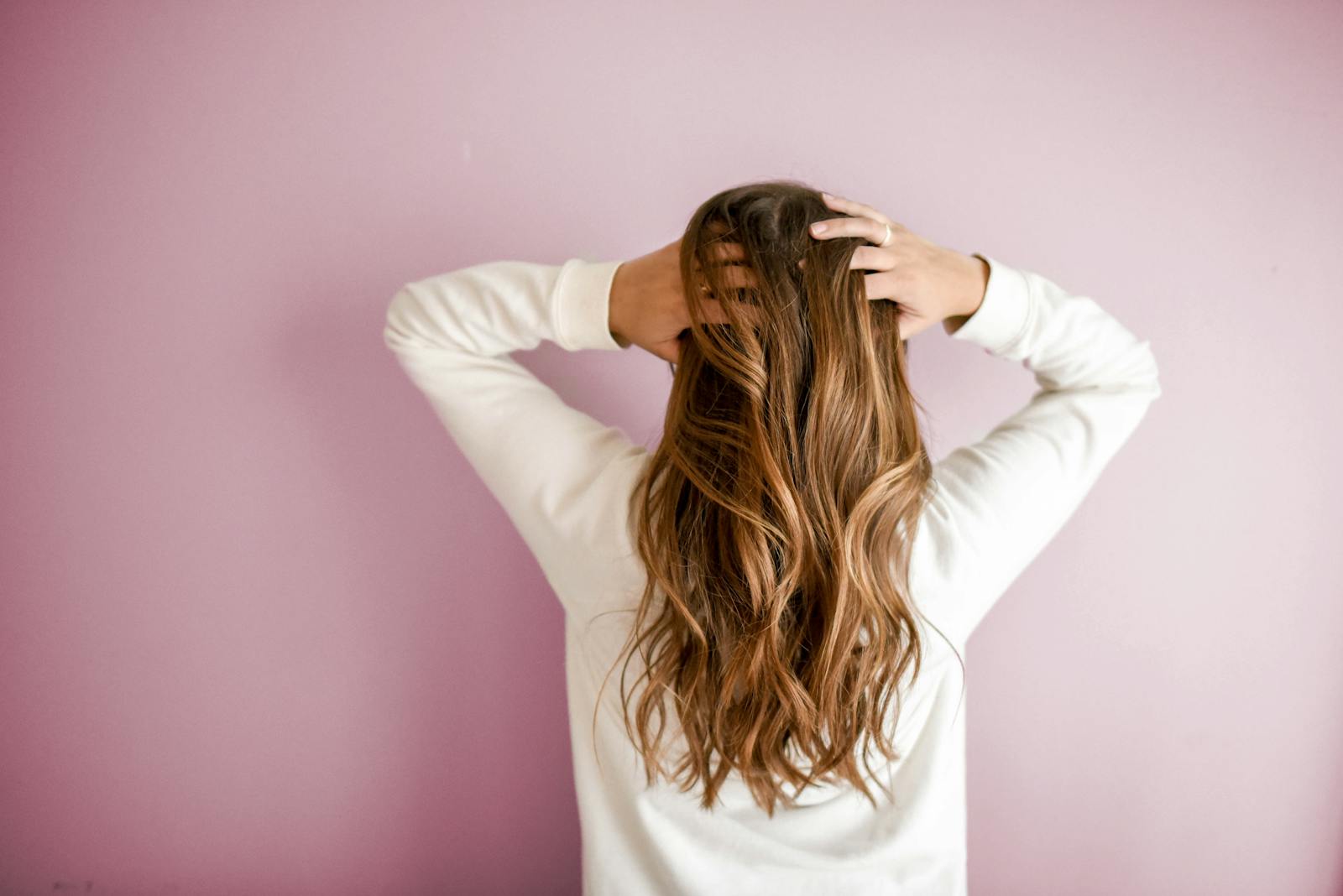 Back view of a woman with elegant long brown hair against a pink wall, showcasing stylish hair design.